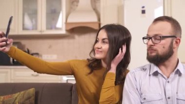 Genç bir esmer ve gözlüklü bir adam stüdyo dairesindeki köşedeki koltukta oturuyorlar; o bir telefon tutuyor ve saçlarını düzelterek, gülümseyerek, yüzlerini çekerek selfie çekiyor.
