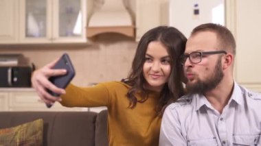 Esmer ve gözlüklü bir adam stüdyo dairesinin köşesinde koltukta oturuyor; bir telefon tutuyor ve selfie çekiyor, gülümsüyor, öpüşüyor, yanağını ısırıyormuş gibi yapıyor, yüzünü çekiyor.