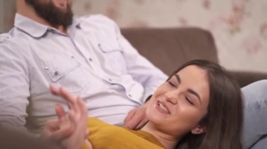 A bearded man in glasses is sitting on the sofa and a pretty brunette lying on his leg, stroking his hand and smiling happily. She touches his nose with her finger