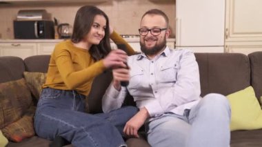 Güzel bir esmer ve gözlüklü sakallı bir adam köşedeki kanepede oturup televizyon izliyor. Adamın elinden uzaktan kumandayı kapmaya çalışıyor. Saklıyor ve kanalları değiştiriyor.