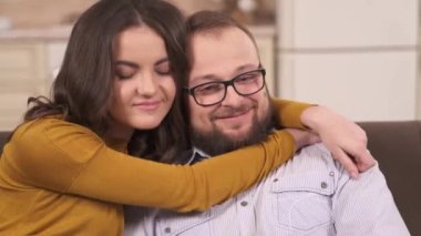 A closeup of a pretty brunette and a bearded man in glasses sitting on the corner sofa and watching TV. She is hugging him and kissing, they are looking in front of them smiling and happy