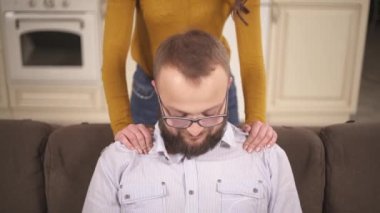 A closeup of a bearded man in glasses sitting on the sofa and woman's hands massaging his shoulders. A pretty brunette bends and puts her head on his shoulder and kisses his cheek