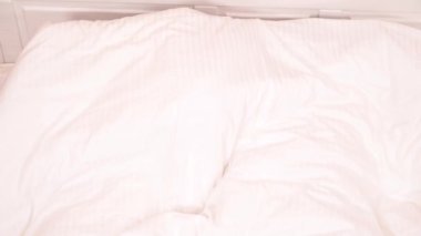 A top view of white blanket on the bed taken away and a pretty brunette with a bearded man lying on the bed; she is smiling, he is yawning and they are hiding under the blanket again