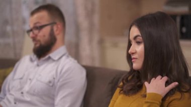 A bearded man and a brunette are sitting on the sofa and making funny faces, laughing and smiling
