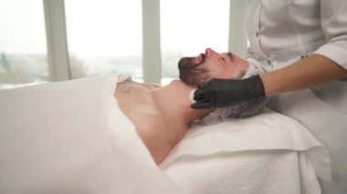 A side view of female hands in black gloves cleansing man's face with cotton sponges in a beauty salon. The man is lying on the couch