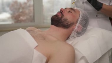 Siyah eldivenli bir kadın eli yağlı bir pipet tutuyor ve bir güzellik salonundaki kanepede yatan adamın alnına döküyor.