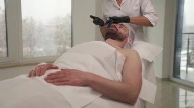 Woman's hands in black gloves applying cream on man's cheekbones, forehead, neck, and massaging his face, neck in a beauty salon. Man is lying on the couch and woman standing at his head