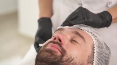 Woman's hands in black gloves rubbing cream with cotton sponges into man's face. Man is lying on the couch in a beauty salon and woman standing at his head