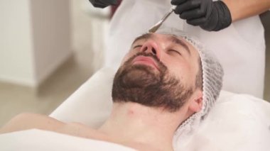 A closeup front view of woman's hands in black gloves applying a mask with a brush on man's forehead in a beauty salon. Man is lying on the couch and woman standing at his head