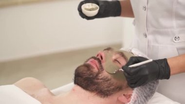 A close-up top view of woman's hands in black gloves applying a mask with a brush on man's cheekbones in a beauty salon. Man is lying on the couch and woman standing at his head