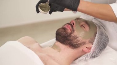 A close-up of woman's hands in black gloves applying a mask with a brush on man's neck in a beauty salon. Man is lying on the couch and woman standing at his head
