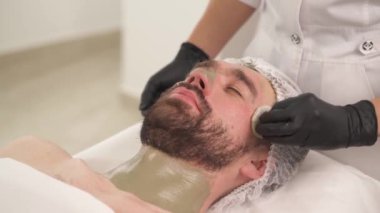Man is lying on the couch and woman standing at his head. The woman cosmetologist continues wiping the mask off the forehead, cheekbones, a nose with cotton pads
