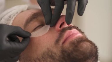 A close-up of woman's hands in gloves applying eye contour pads under the right eye on man's face and adjusting it. The man is lying on the couch in a beauty salon and woman standing at his head