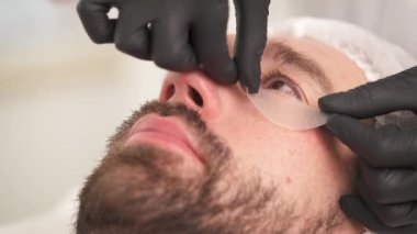 A close-up of woman's hands in gloves applying eye contour pads under the left eye on man's face and adjusting it. The man is lying on the couch in a beauty salon and woman standing at his head