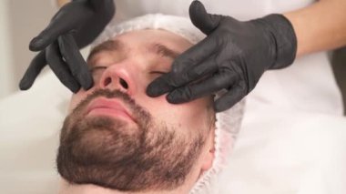 A closeup of woman's hands in gloves pressing and adjusting the skin with applied eye contour pads on man's face. The man is lying on the couch in a beauty salon and woman standing at his head