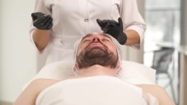 A closeup front view of woman's hands in black gloves applying cream on man's face in a beauty salon. The man is lying on the couch and the cosmetologist is standing at his head