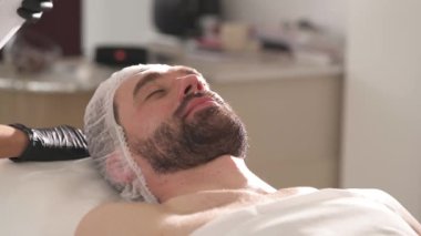 A close-up side view of man's face in a beautician cap lying on the couch. The white spray is sprinkled above him