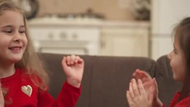 Two happy little girls are sitting on the sofa and playing pat-a-cake