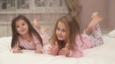 Cute little girls in pajamas are laying on stomach on the bed , moving crossed legs and talking.