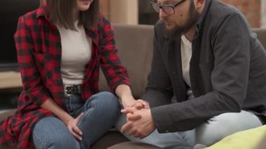 Young bearded man in glasses is upset and disappointed. He is sitting on the sofa with his girlfriend. She carefully takes his hand and tries to support him in every possible way.