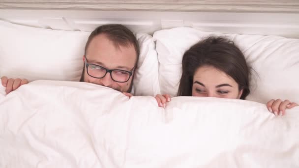 若いカップルはベッドの上に横たわって毛布の外を見る 彼らはカメラを見て 互いを見て 点滅し — ストック動画