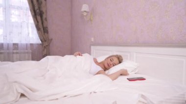 A pretty blonde sleeping in a big bed with white linen, stretching and waking up, taking her phone, looks at it and jumps in the bed