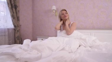 A charming blonde sitting in bed, massaging her neck, stretching arms in front of her, rotating wrists and stretching arms to the sides and rising hands up