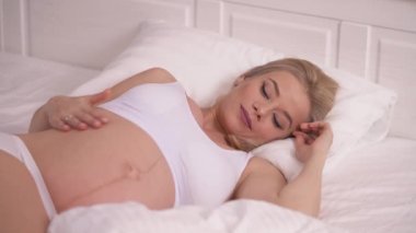 A pretty pregnant blonde lying in bed and caressing her belly gently; she stops and smiles and touches it tenderly