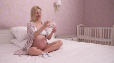 A pregnant blonde in a gown sitting cross-legged on the bed and holding a pink headband with decorations; she looks at it and puts it on her belly with tenderness