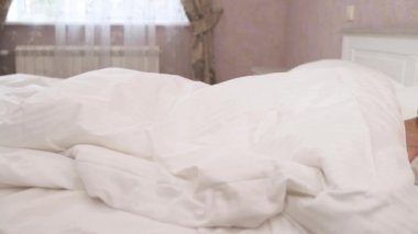 The white blanket on the bed, covering a sleeping blonde. The camera moves from left to right and stops at a pretty blonde's face lying on the bed and sleeping peacefully