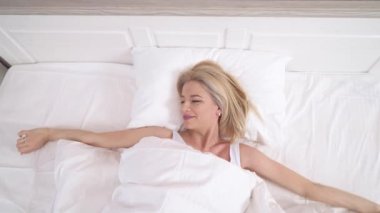 A pretty blonde lying in bed with white linen, stretching her arms to the sides and above her, waking up