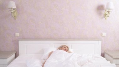 A front view of a pretty blonde sleeping in bed, sitting on it, stretching her arms and tumbling hair