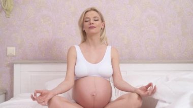 A close-up of a pregnant woman sitting cross-legged on the bed, keeping her hands on her knees and doing yoga exercise at home. The camera moves backwards