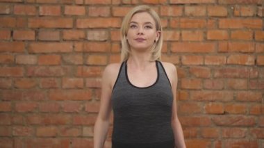 A pregnant blonde in dark tank top standing, rotating shoulders, holding her arms bent, and turning the body. Pregnant woman exercising against a brick wall in the background