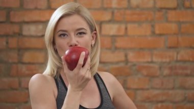A close-up of a charming blonde in dark tank top standing against the brick wall and biting a big red apple and chewing it
