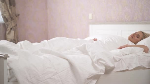 床上有一条白色的毯子床底下睡着一个漂亮的金发美女她醒来 坐在床上 转向窗户 微笑着举起她的手 照像机 — 图库视频影像