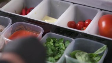 Plastic containers with sauce, parsley, spinach. A plastic lid covers another container and hands in black gloves open a lid and take cherry tomatoes from the container