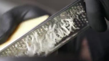 A close-up of hands in black gloves holding a grater and a piece of cheese and grating it. Low angle view