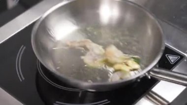 A frying pan with garlic and spinach and oil on the stove, then a hand in black glove adds more ingredients: shrimps, tomatoes, and sprinkles the mixture with oil, shakes it and puts a pan back