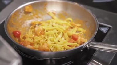 Sebze soslu erişteli bir tava, karides; bir el onları sallıyor ve kaşıkla su ekliyor, onları mutfak cımbızıyla karıştırıyor, karışımı sallıyor ve tavayı ocağın üzerine koyuyor.