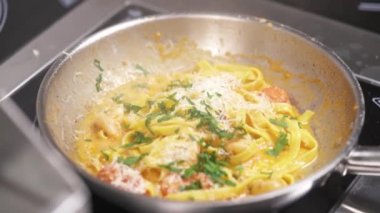 Noodles in sauce of vegetables, shrimps sprinkled with grated cheeses on top sizzle in a frying pan, a hand adds greens and mixes it with culinary tweezers, adds water and shakes the pan