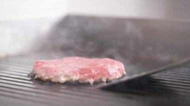 A close-up of a burger frying on the grill and a hand in black glove turning it over with a kitchen paddle