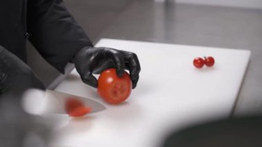 Beyaz bir tahta, yanında da domates, domates ve bıçak var. Profesyonel mutfakta siyah eldivenli eller beyaz tahtada domates dilimliyor.