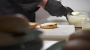 Toasted halves of buns lying on the white board on the table and hands in black gloves put white sauce on one half and salad over it