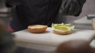 Two hands in black gloves putting pickles, a burger with melted cheese, tomatoes, slices of pickled onions on a toasted half of the bun with lettuce. The second half lies near it