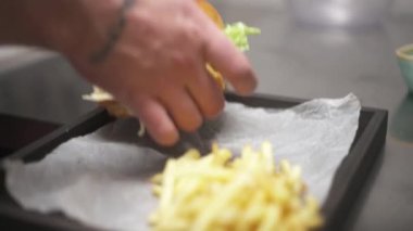 Hands putting a finished burger with a stick in the middle next to French-fries lying on the tray. The camera moves bottom up