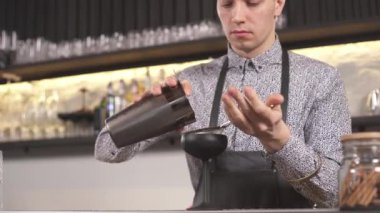 A bartender standing at the bar counter, he takes a shaker with prepared cocktail and puts a black tumbler on the counter, puts the sieve on the shaker and pours the cocktail into the tumbler