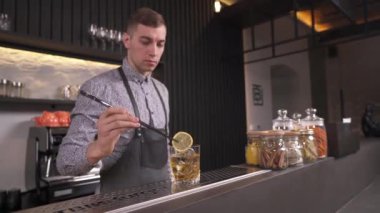 Bir barmen tezgahın üzerinde duran bir bardak viskinin içine uzun cımbızla bir dilim limon koyuyor. Tezgahın arkasında bir barmen duruyor.