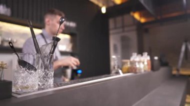 A close-up of cocktail utensils standing in glasses on the bar counter, then a bar tender takes a tumbler twisting it twice and putting on the counter in front of him