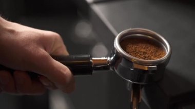 A close-up of a hand holding a portafilter with ground coffee on the edge of the table, another hand presses it with a tamper. Coffee making in a bar